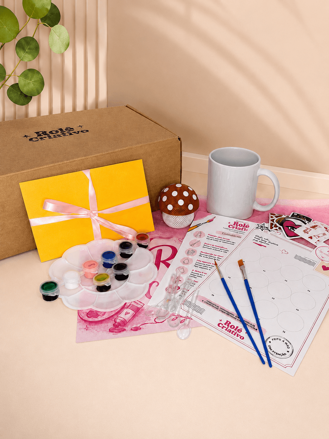 Caixa do Rolê Criativo aberta, com tintas, pincéis, caneca, cartinha e o kit de pintura